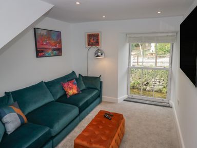 A living room with a teal sofa orange ottoman floor lamp paintings and a window showing an outside view at Rose Cottage in Ambleside