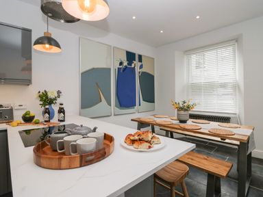 A kitchen with a white countertop holding a wooden tray with teapot and cups and a plate of baked goods next to a wooden dining table with placemats and a plant at Rose Cottage in Ambleside