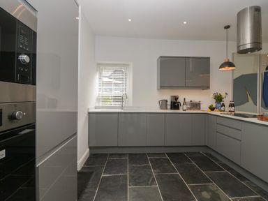 A kitchen with grey cabinets, black tiled floor, built-in oven, sink under window, coffee maker, kettle, toaster, bottles, and hanging light at Rose Cottage in Ambleside
