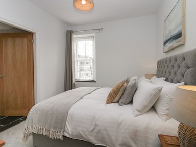 A bedroom with a gray upholstered bed with multiple pillows beside a window with curtains at Rose Cottage in Ambleside