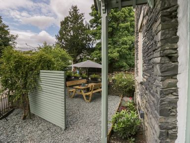 An outdoor seating area with wooden benches and table under an umbrella surrounded by plants and a stone wall at Rose Cottage in Ambleside