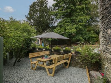 An outdoor seating area with a wooden picnic table and benches with an umbrella surrounded by plants and stone walls at Rose Cottage in Ambleside
