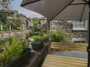 An outdoor seating area with a wooden bench and large umbrella overlooking a stone-lined river and old stone buildings at Rose Cottage in Ambleside