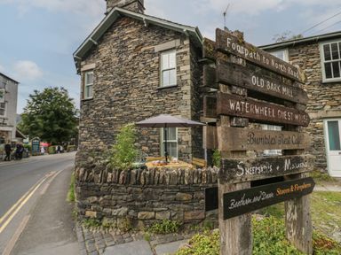 A stone building beside a road with a wooden signpost showing local destinations and a small outdoor seating area with a table umbrella