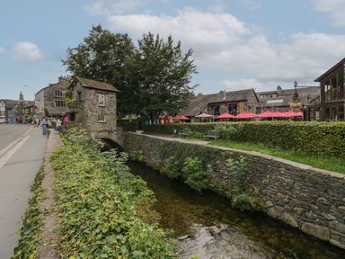 A stone bridge with a small building over a stream and outdoor seating with red umbrellas at Rose Cottage in Ambleside