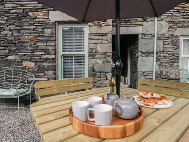An outdoor wooden table with teapot cups wine bottle and pastries under an umbrella by a stone wall at Rose Cottage in Ambleside