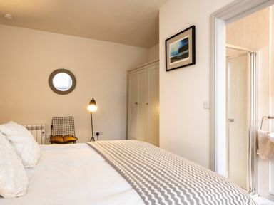 A bedroom with a bed a white wardrobe a chair with a cushion a floor lamp a round mirror and an en suite bathroom at The Lookout in Polzeath