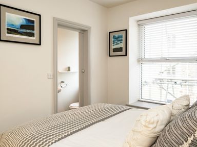 A bedroom with a bed and pillows next to a window with blinds and a door leading to a bathroom at The Lookout in Polzeath