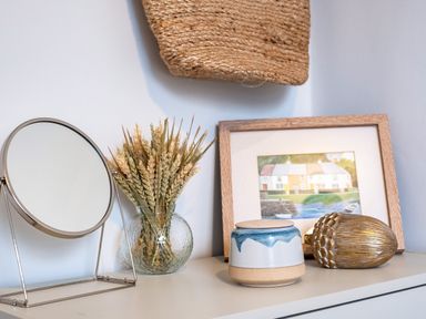 A table with a round mirror a glass vase with wheat stalks a ceramic jar a gold acorn decoration and a framed picture at The Lookout in Polzeath