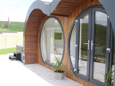 A wooden building exterior with large circular windows and glass doors and a potted plant on the patio at Creuddyn in Llanfihangel-y-Creuddyn near Aberystwyth