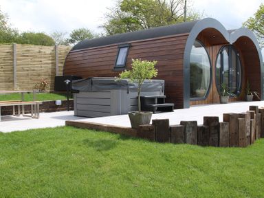 A wooden curved cabin with potted plants on a patio and a covered hot tub in a fenced garden at Creuddyn Llanfihangel-y-Creuddyn near Aberystwyth