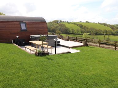 A wooden cabin with a hot tub and outdoor seating on a patio overlooking green hills and trees at Creuddyn in Llanfihangel-y-Creuddyn near Aberystwyth