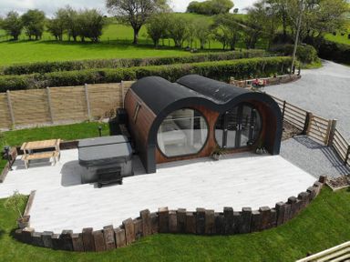 A small curved wooden cabin with large round window and doors on a white tiled patio next to a hot tub and picnic table at Creuddyn in Llanfihangel-y-Creuddyn near Aberystwyth