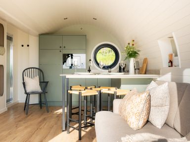 A compact kitchen and dining area with a green cabinet unit a round window stools a black chair and a sofa with cushions at Creuddyn in Llanfihangel-y-Creuddyn near Aberystwyth