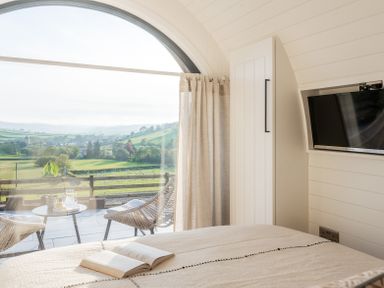 A bedroom with a bed an open book a wall-mounted TV and a balcony with chairs and a table overlooking green fields at Creuddyn in Llanfihangel-y-Creuddyn near Aberystwyth