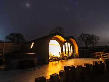 A modern curved cabin with illuminated interior and two chairs on the wooden deck under a starry sky at Creuddyn in Llanfihangel-y-Creuddyn near Aberystwyth