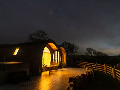 A curved wooden cabin with lit interior and two chairs on a tiled patio at night surrounded by a wooden fence and trees at Creuddyn in Llanfihangel-y-Creuddyn near Aberystwyth