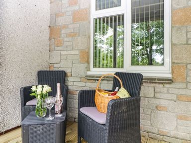 A patio with two black wicker chairs a small black table with white roses a bottle and two glasses and a basket with bottles by a window at 17 Valley Lodges in Gunnislake