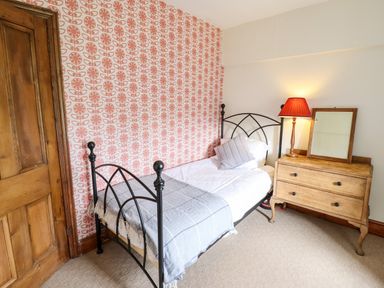 A bedroom with a single bed a wooden dresser with a mirror and a lamp a wooden door and patterned wallpaper at Gwengraig in Brithdir near Dolgellau
