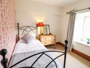 A bedroom with a single bed a wooden chest of drawers with a lamp and mirror and a window with curtains at Gwengraig in Brithdir near Dolgellau