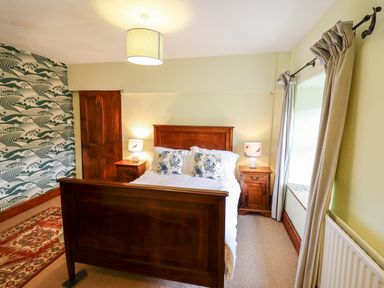 A bedroom with a wooden bed, two bedside tables with lamps, patterned wallpaper, and beige curtains at Gwengraig in Brithdir near Dolgellau