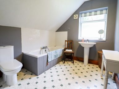 A bathroom with a toilet a bathtub with a towel a wooden chair a pedestal sink a window and a white table at Gwengraig in Brithdir near Dolgellau