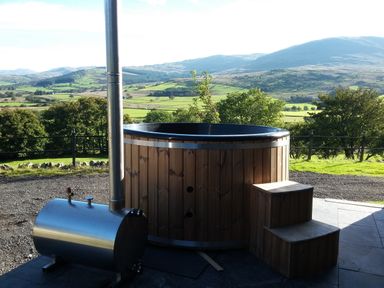 A wooden hot tub with metal stove and steps outside with a view of green fields and hills at Gwengraig in Brithdir near Dolgellau