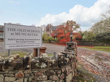 A stone wall with a sign for The Old School at Muncaster and a driveway leading to a building with trees in Ravenglass near Holmrook