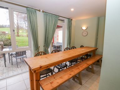 A dining room with wooden table and bench seating next to large windows and green curtains at Old School Muncaster House Ravenglass near Holmrook