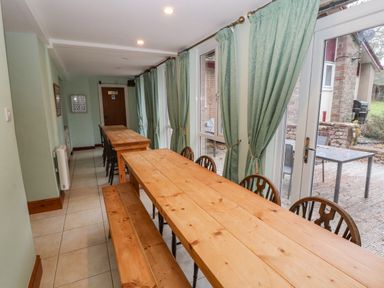 A dining area with wooden tables, benches, chairs, green curtains, tiled floor, and glass doors opening to an outdoor patio at Old School Muncaster House Ravenglass near Holmrook
