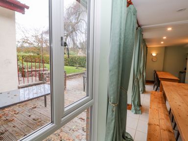 A dining area with long wooden tables and benches beside windows with green curtains and a view of a garden patio at Old School Muncaster House Ravenglass near Holmrook