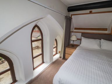 A bedroom with a bed a bedside table a lamp and arched windows at Old School Muncaster House in Ravenglass near Holmrook