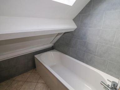 A bathroom with a white bathtub under a sloped ceiling and gray tiled walls at Old School Muncaster House Ravenglass near Holmrook