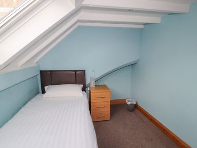 A single bed with pillow next to a small wooden drawer in a room with blue walls and slanted ceiling at Old School Muncaster House Ravenglass near Holmrook