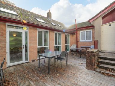 An outdoor patio area with tables and chairs on a wooden deck outside a brick building at Old School Muncaster House in Ravenglass near Holmrook