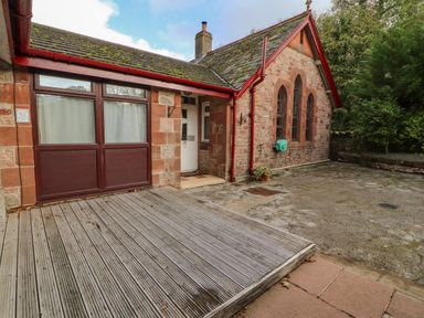 An exterior view of a stone building with wooden deck and paved ground at Old School Muncaster House in Ravenglass near Holmrook