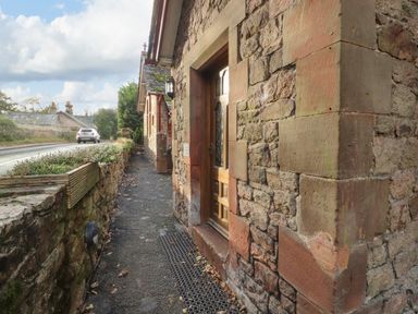 A stone building with a wooden door along a narrow pathway next to a road at Old School Muncaster House Ravenglass near Holmrook