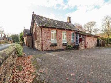 A stone building with a sloped roof and windows by a paved road at Old School Muncaster House Ravenglass near Holmrook