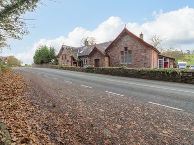 A stone house with red trim along a road with fallen leaves and a stone wall at Old School Muncaster House in Ravenglass near Holmrook