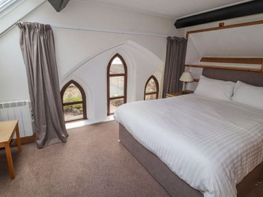 A bedroom with a large bed beside three arched windows and a small wooden table at Old School Muncaster House Ravenglass near Holmrook