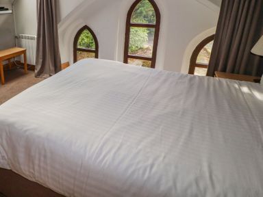 A bed with white linens beside arched windows with curtains and a wooden desk at Old School Muncaster House Ravenglass near Holmrook
