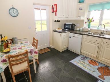 A kitchen with a dining table and chairs a door a clock white cabinets a microwave a dishwasher a sink and a window with flowers on the sill at North Studdock Cottage in Angle near Pembroke