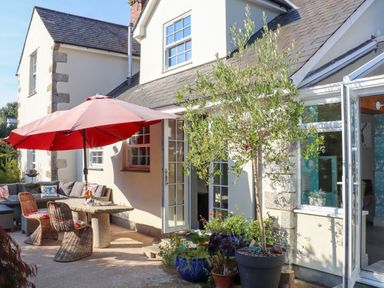 An outdoor patio with a table and chairs under a large red umbrella and potted plants near an open door at Lower Gadles Burnthouse near Falmouth