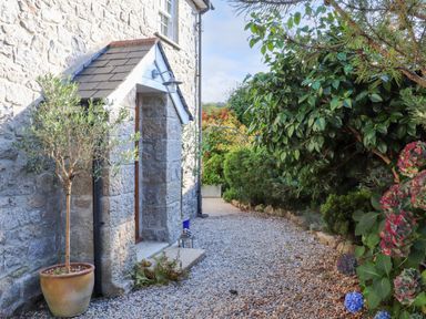 A stone house entrance with a potted tree and a gravel pathway lined with bushes at Lower Gadles in Burnthouse near Falmouth