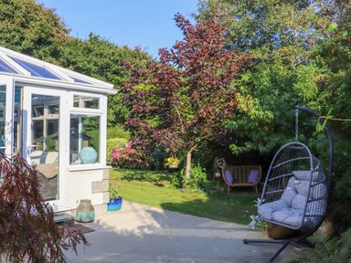 A garden with a hanging chair and a bench near trees and a conservatory at Lower Gadles Burnthouse near Falmouth