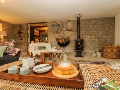 A living room with a stone wall and wood-burning stove with a tray of tea and cake on a wicker table at Lower Gadles Burnthouse near Falmouth