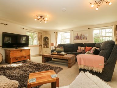 A living room with a large dark sofa a wooden coffee table a wooden TV stand with a television and windows with curtains at Lower Gadles in Burnthouse near Falmouth