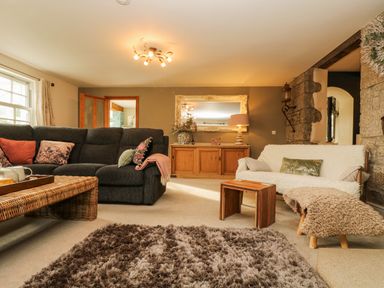 A living room with a dark sofa a wicker coffee table a wooden sideboard a large mirror and a light-colored armchair with a sheepskin throw at Lower Gadles Burnthouse near Falmouth
