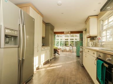 A kitchen with stainless steel refrigerator dishwasher oven and sink leading to a dining area with white chairs and large windows at Lower Gadles Burnthouse near Falmouth