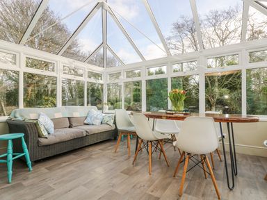 A conservatory with gray sofa and white chairs around wooden table with flowers at Lower Gadles Burnthouse near Falmouth